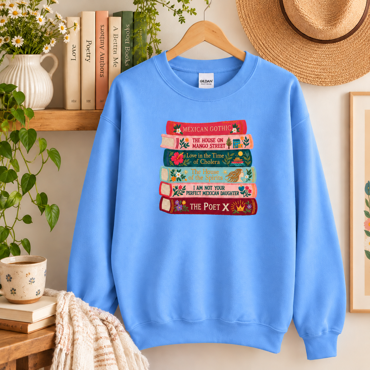Carolina Blue Gildan crewneck sweatshirt with colorful embroidered Latina literature books hanging on a wooden hanger.