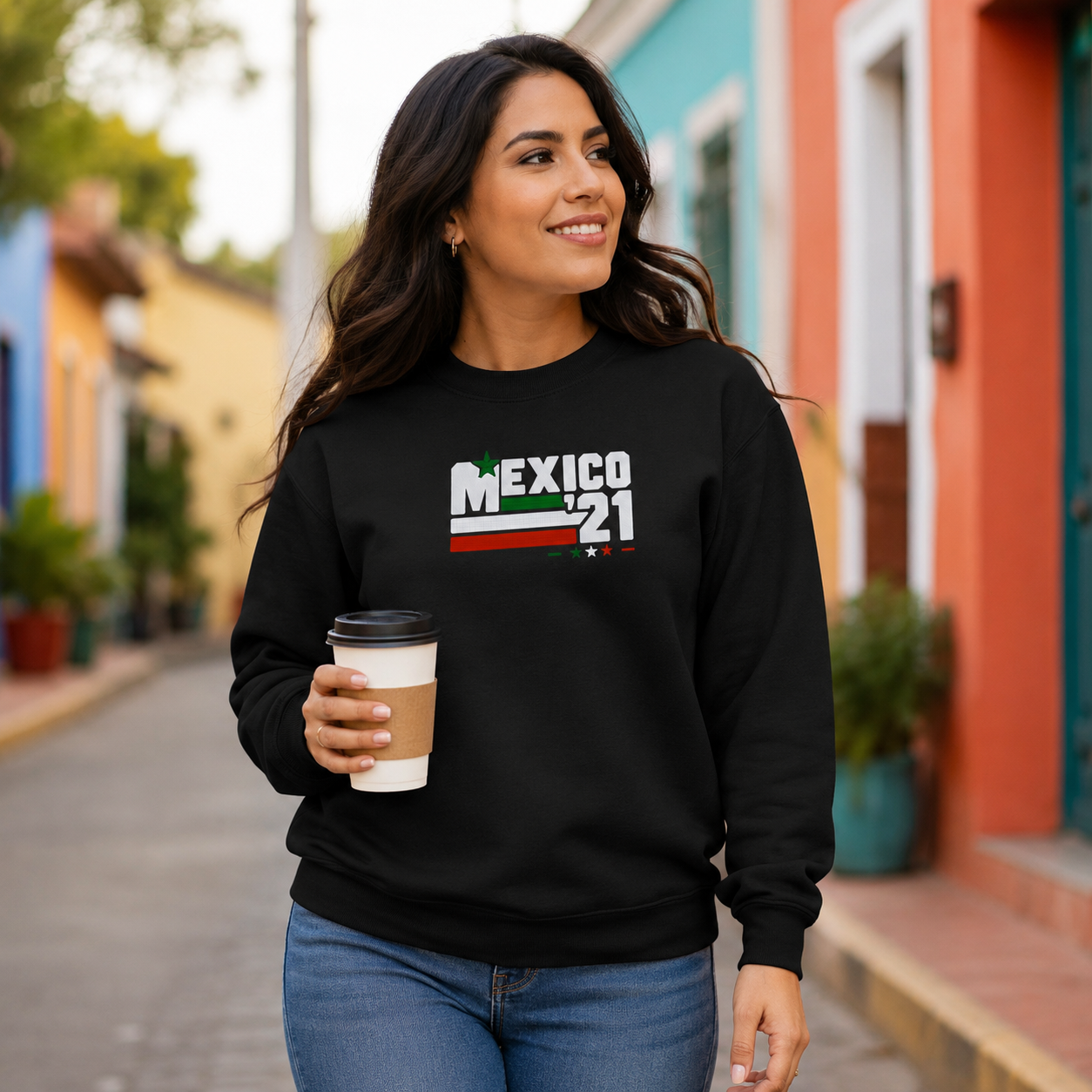 Woman wearing a black Gildan Heavy Blend crewneck sweatshirt featuring Embroiderzilla's retro Mexico '21 embroidered design while holding a coffee and walking through a colorful neighborhood.