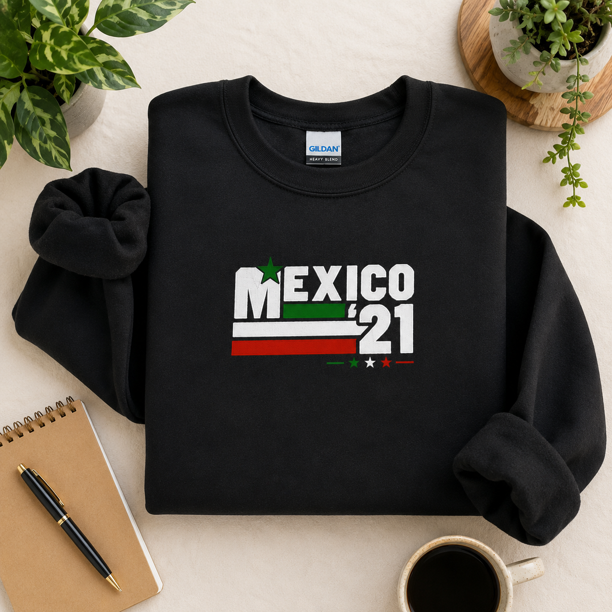 Black Gildan Heavy Blend crewneck sweatshirt featuring Embroiderzilla's retro Mexico '21 embroidered design styled in a cozy flat lay with a notebook, coffee, and plants.