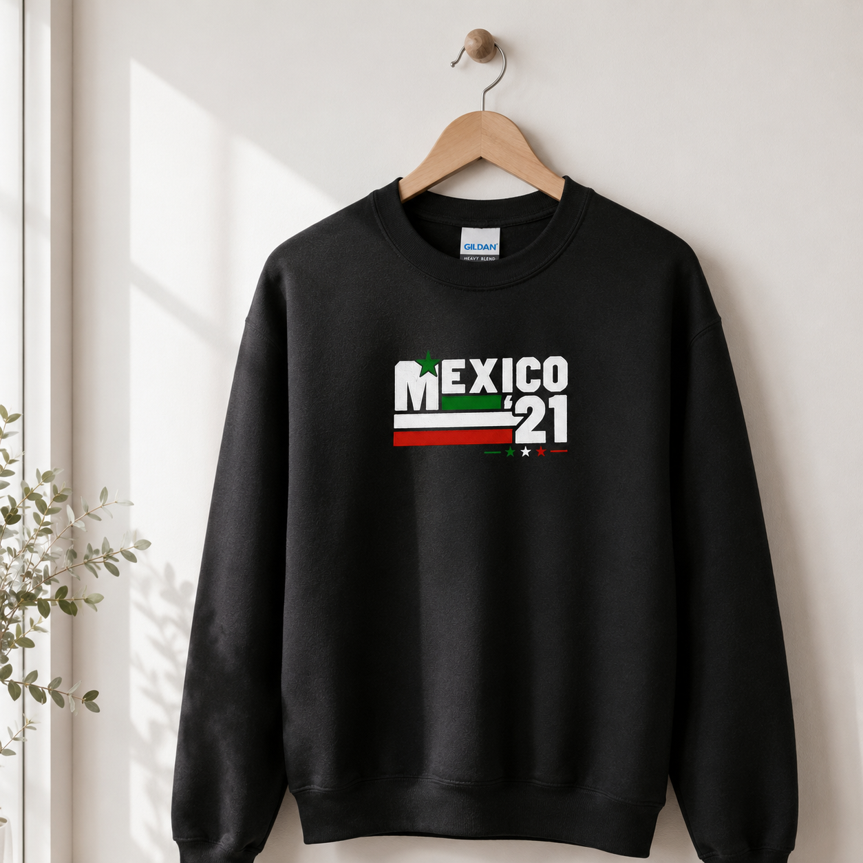 Black Gildan Heavy Blend crewneck sweatshirt featuring Embroiderzilla's retro Mexico '21 embroidered design hanging on a wooden hanger in natural window light.