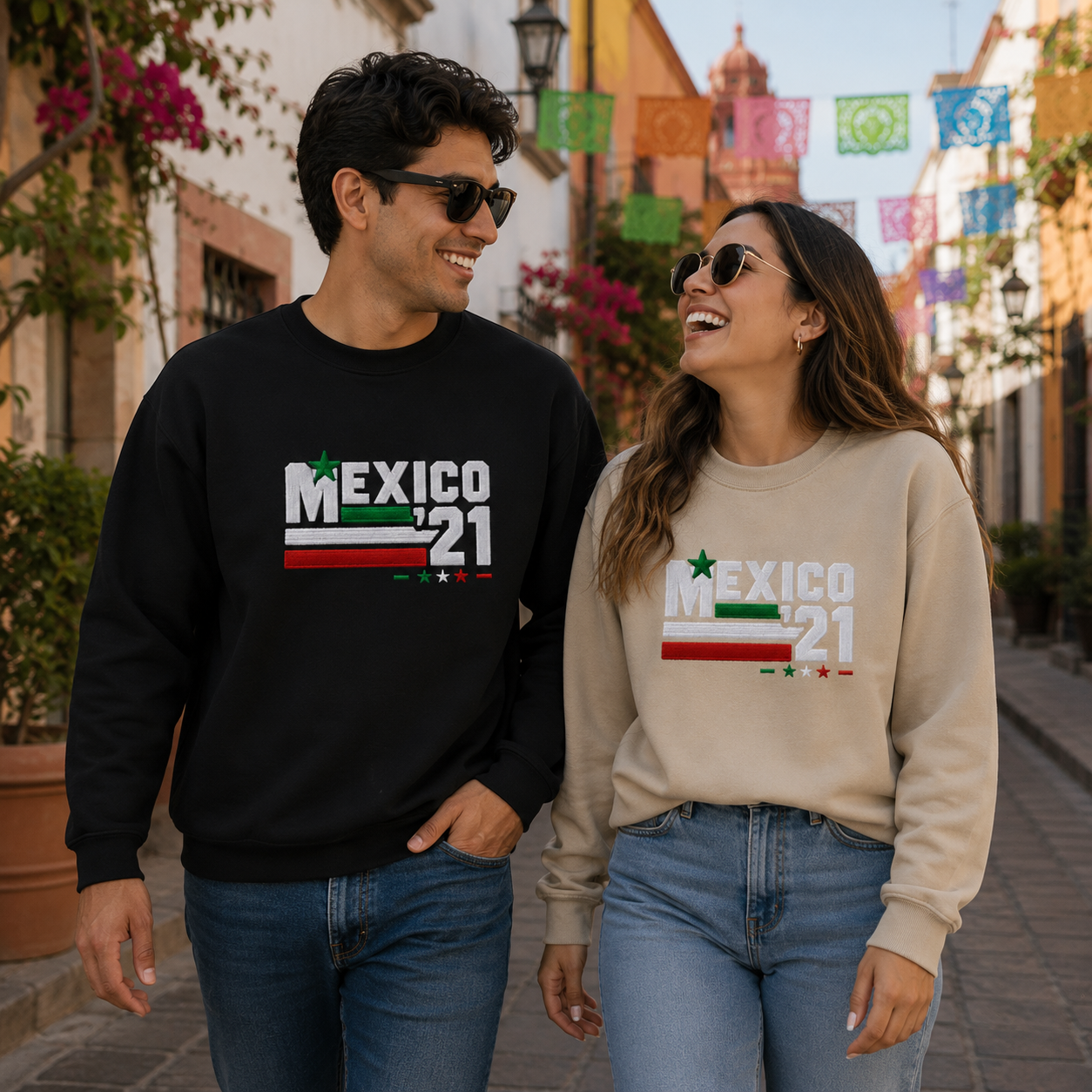 Couple wearing black and sand Gildan Heavy Blend crewneck sweatshirts featuring Embroiderzilla's retro Mexico '21 embroidered design while walking through a colorful historic street.
