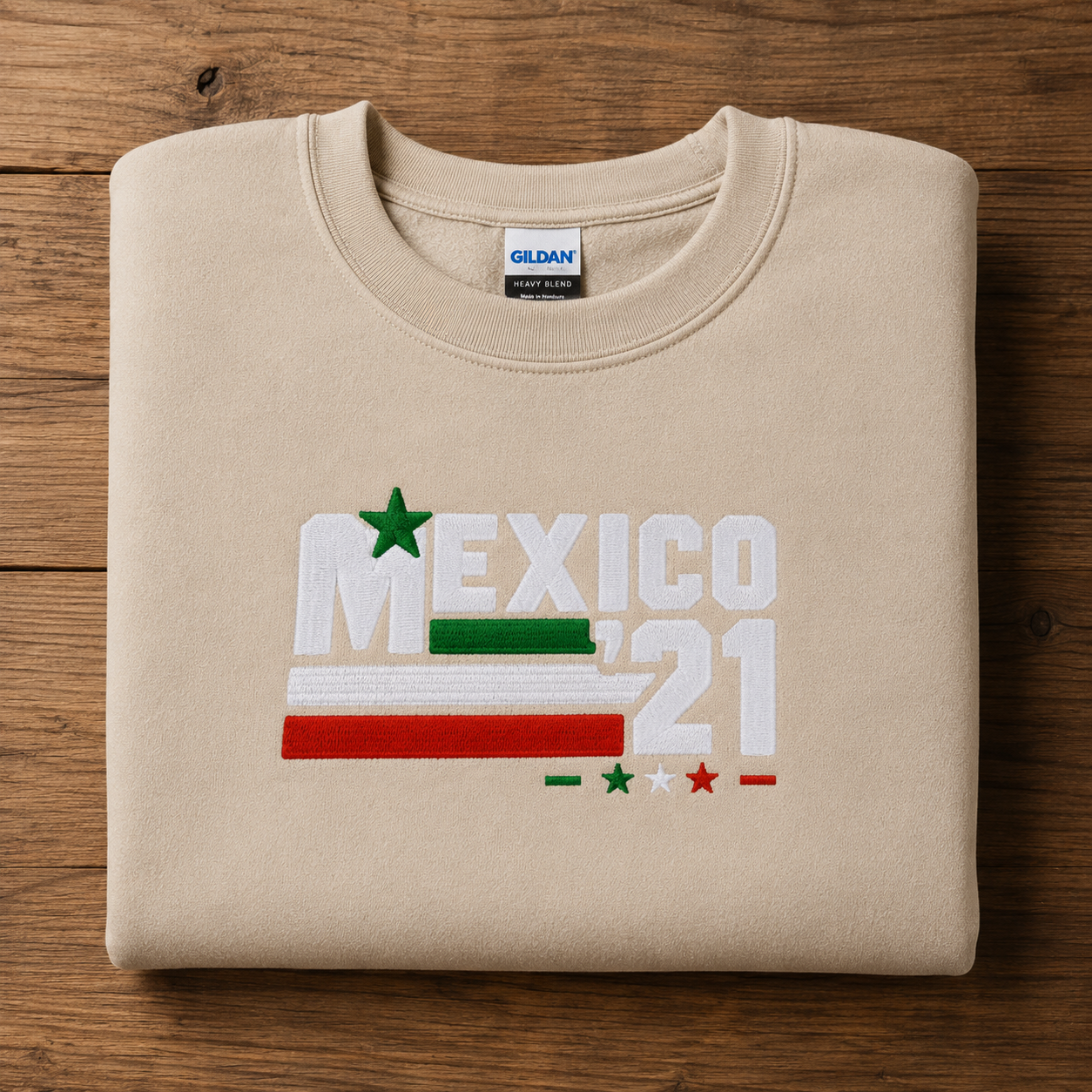 Folded sand Gildan Heavy Blend crewneck sweatshirt featuring Embroiderzilla's retro Mexico '21 embroidered design displayed on a wooden tabletop.